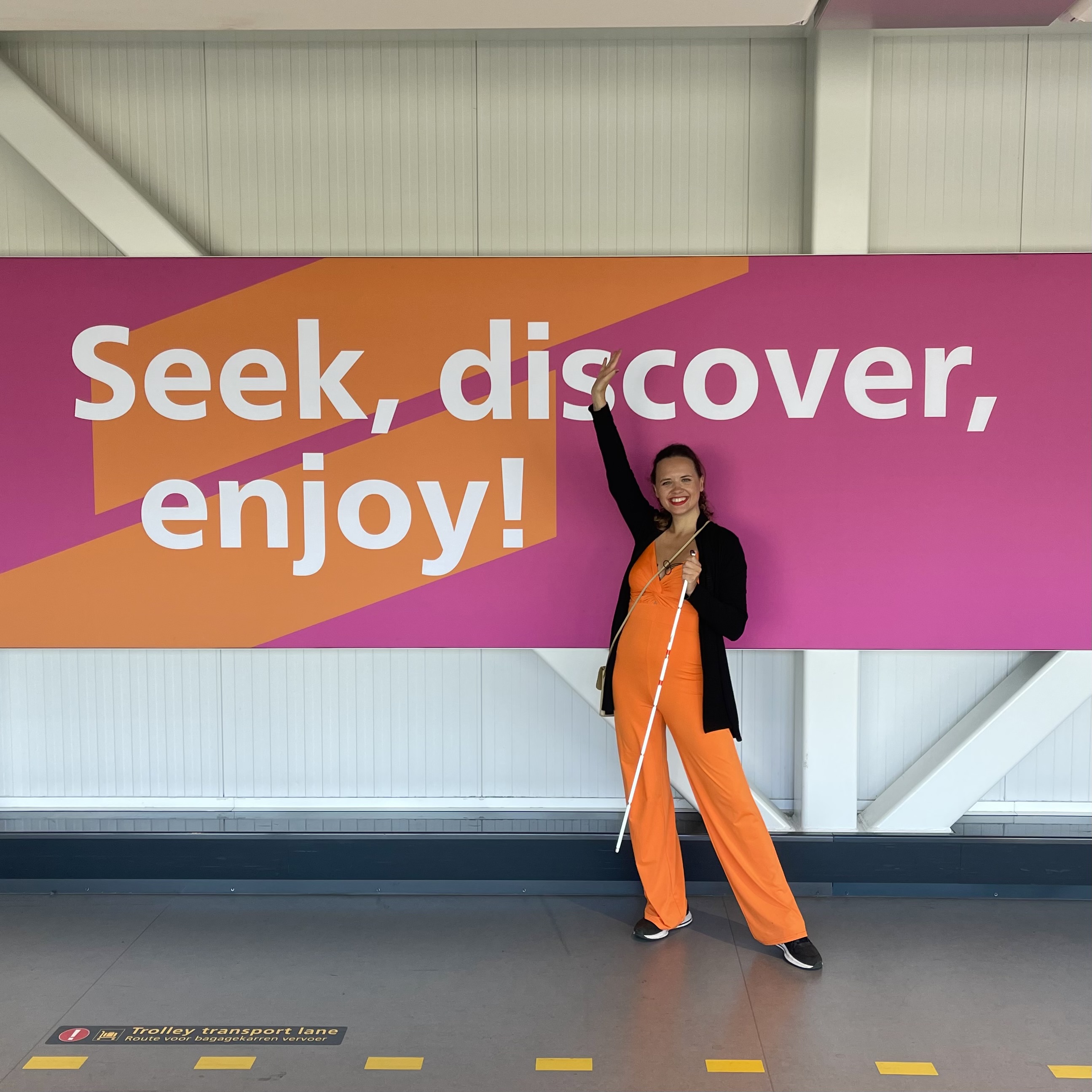 “Seek, Discover, Enjoy:” Marie Elise, wearing a bright orange jumpsuit and a long black sweater, is standing in front of a large sign at Amsterdam Airport that reads: “Seek, Discover, Enjoy.” She has her right arm raised in the air, and in her left hand, she’s holding her symbol cane. The letters on the board are white, set against an orange and pink background.